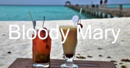 Auf dem Bild steht "Bloody Mary" in weißer Schrift, während zwei Cocktails auf einem Tisch platziert sind, im Hintergrund sind ein Strand und das Meer zu sehen.